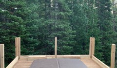 Covered hot tub built into wooden deck with partially-constructed railings overlooking forest